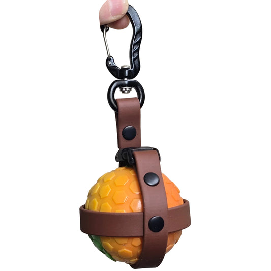 A detailed close-up of a handcrafted brown BioThane ball holder. It is secured by a durable, high-strength black aluminum carabiner. The BioThane strap holds a bright orange tennis ball (for demonstration), showing a waterproof and easy-to-clean way to carry toys hands-free. 