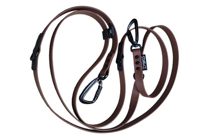 A brown BioThane hands-free adjustable dog leash (3m) arranged in a loop on a white background. Features dual premium black auto-lock carabiners, tactical D-rings, black rivets, and a black DoggoZ brand tag.